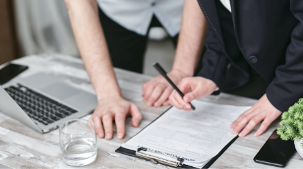 Zwei Personen arbeiten zusammen an einem Schreibtisch, eine Person unterschreibt ein Dokument auf einem Klemmbrett